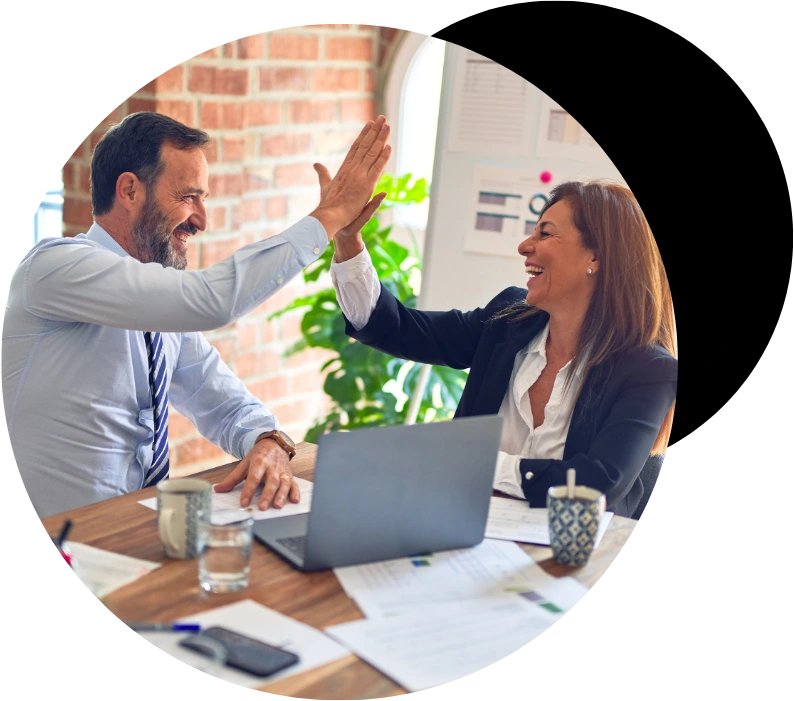 A smiling male executive in a blue shirt and tie giving a high five to a laughing female business partner in a black blazer at a wooden office desk equipped with a laptop, documents, and coffee mugs against a brick background.