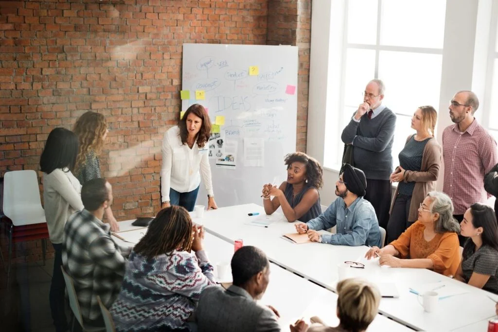 An indoor corporate strategy meeting showing a diverse group of employees seated around long white tables in a rustic brick room, actively listening to a woman standing at the front presenting ideas on a whiteboard filled with diagrams and sticky notes.