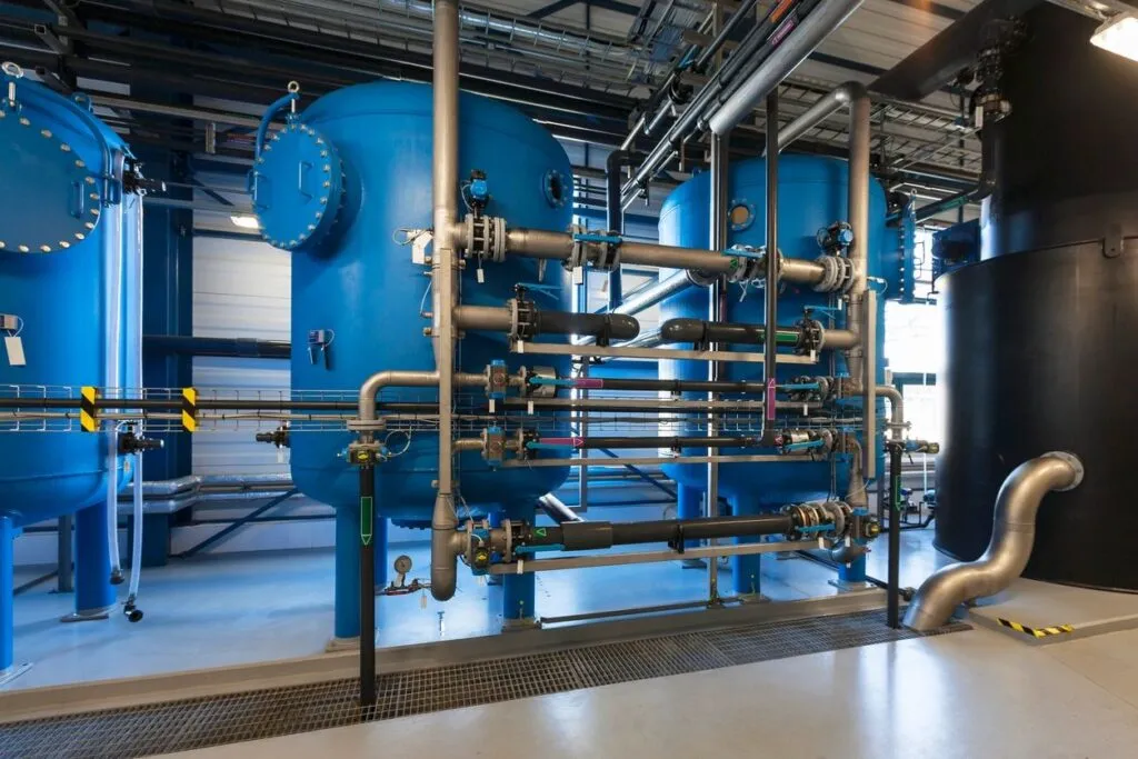 Wide industrial shot of an indoor water purification facility featuring a sequence of large, bright blue vertical pressure vessel tanks interconnected by complex stainless steel pipelines, control valves, and electrical conduits.